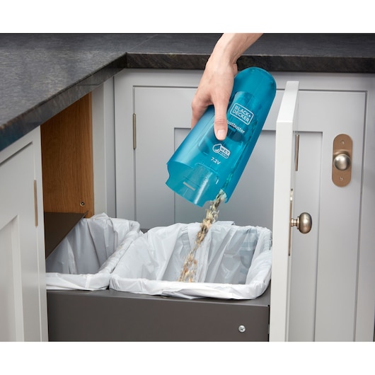 A person is emptying the dust container of a blue BLACK+DECKER handheld vacuum cleaner into a trash bin inside a kitchen cabinet.