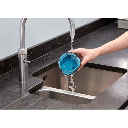 A person rinsing a blue vacuum cleaner filter under a kitchen faucet, with water flowing into a stainless steel sink.