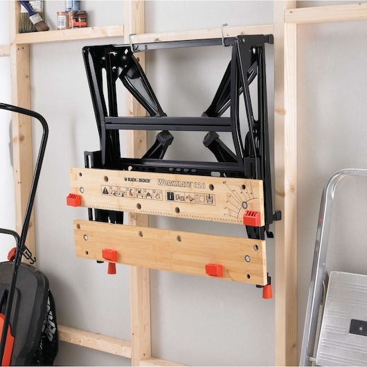 BLACK+DECKER WM626 folding workbench with wooden top and orange clamps, wall-mounted in a garage.
